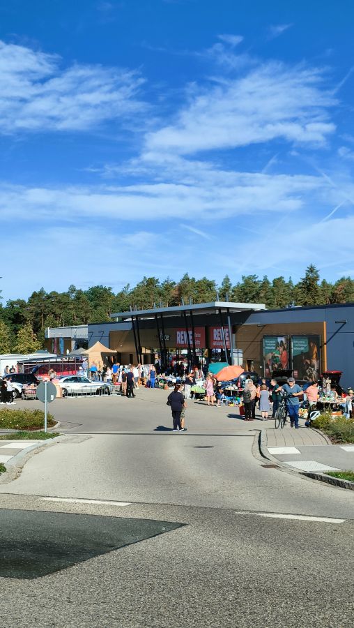 Hochformat-Uebersicht des Flohmarkts am REWE in Rednitzhembach: ueber dem Marktgebaeude viel blauer Himmel, davor Staende, Besucher und ein Radfahrer auf der Marktstrasse.