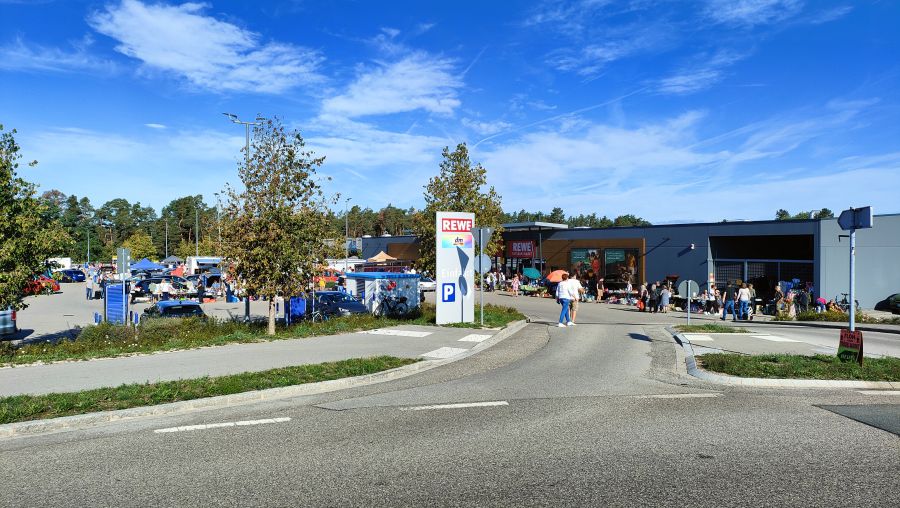 Uebersicht des Flohmarkts am REWE in Rednitzhembach von der Kreuzung aus: REWE-Stele, Staende auf dem Parkplatz und entlang des Gebaeudes, Besucher und ein Flohmarkt-Aufsteller am Strassenrand.