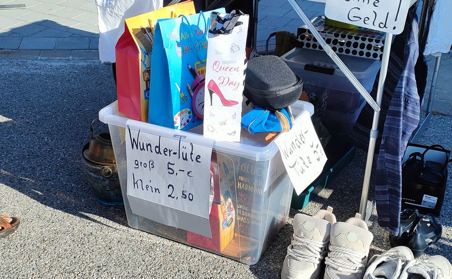 Durchsichtige Kunststoffbox mit Preisschild „Wunder-Tüte groß 5,-€ / klein 2,50" auf dem Flohmarkt am REWE in Rednitzhembach, obenauf bunte Geschenktüten; daneben ein zweites Schild „Wunder-Tüte 3€".