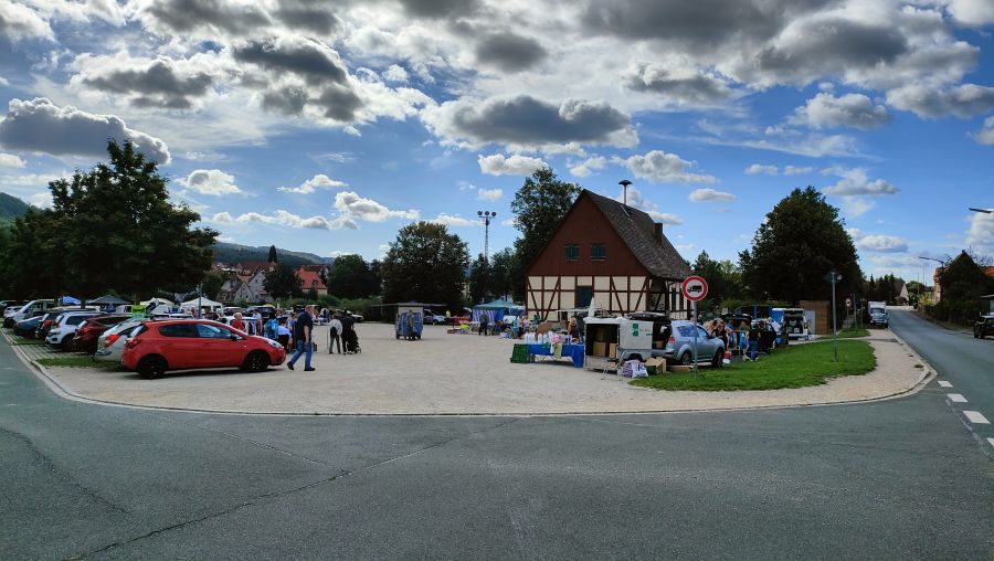 Weiter Blick ueber den Festplatz am Weiher in Schnaittach mit Troedelmarkt: eine Kiesflaeche, ein rotbraunes Fachwerkhaus, parkende Autos und Marktstaende unter dramatisch bewoelktem Himmel, im Hintergrund bewaldete Huegel.