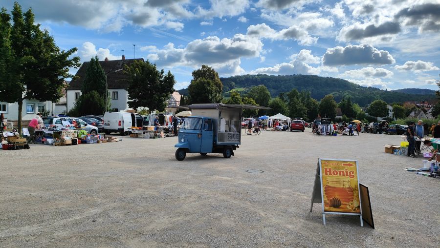Troedelmarkt auf dem Festplatz am Weiher in Schnaittach: in der Mitte eine blaue Piaggio Ape als Eis-Verkaufswagen, daneben ein gelbes Honig-Schild, ringsum Staende, Autos und Besucher, im Hintergrund Huegel.