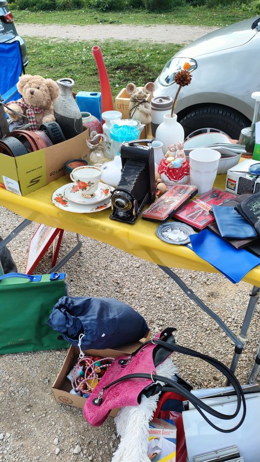 Troedeltisch mit gelber Decke auf dem Markt in Schnaittach: ein Teddybaer, Vasen, eine alte Balgen-Klappkamera, eine rosengemusterte Teetasse, Lederguertel in einer Kiste, DVDs und Geldboersen; darunter am Boden pinke Cowboystiefel in einem Karton.