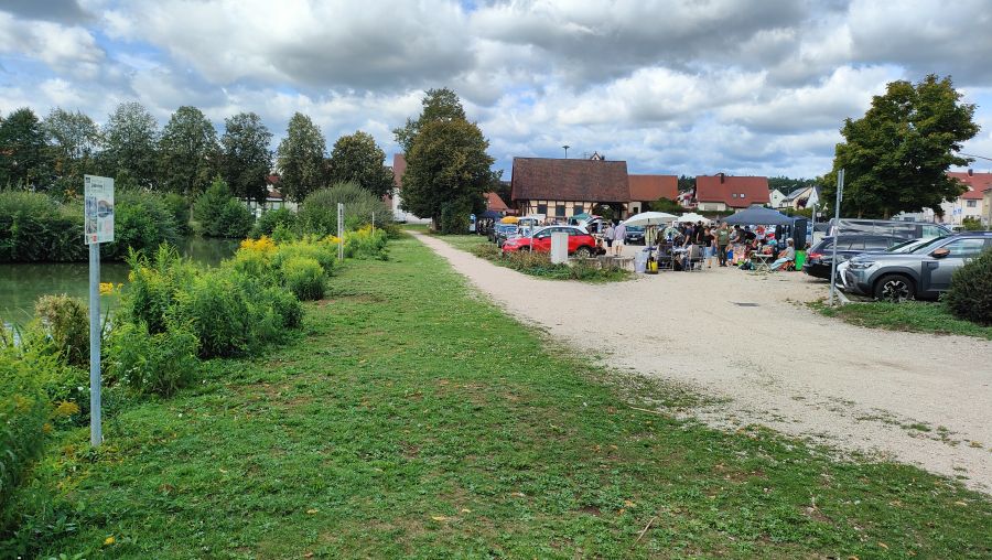 Blick vom Weiher auf den Festplatz in Schnaittach: links der von Schilf und Herbstblumen gesaeumte Teich mit einem Lehrpfad-Schild, in der Mitte ein Kiesweg, rechts der Troedelmarkt mit Staenden, Pavillons, Autos und einem Fachwerkhaus.
